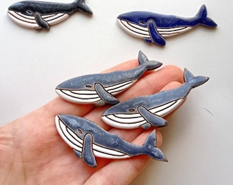 May include: A collection of ceramic whale figurines in various shades of blue, gray, and black. Each whale has a white belly and is hand-painted with detailed fins and tail. Some whales are held in a hand, while others are arranged on a white surface.