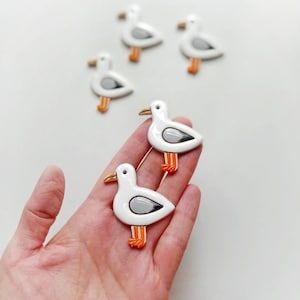 Op de afbeelding: Witte zeemeeuwspelden met grijze en oranje accenten. De spelden hebben een glanzende afwerking en zijn geplaatst op een witte achtergrond. Een speels accessoire voor kleding of tassen.