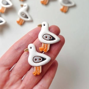 Może przedstawiać: Białe guziki w kształcie mewy z szarymi i czarnymi detalami oraz pomarańczowymi stopami. Guziki mają błyszczące wykończenie i są ułożone na białej powierzchni. Dwa guziki trzymane są w dłoni, a inne rozrzucone w tle.