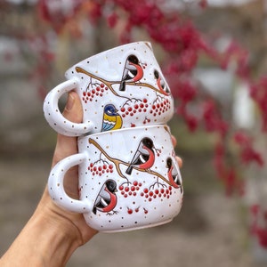 Puede incluir: Dos tazas de cerámica blanca con asas, apiladas una encima de la otra. Las tazas están decoradas con un fondo blanco, puntos dorados y un patrón de pájaros rojos y azules posados ​​en ramas con bayas rojas.