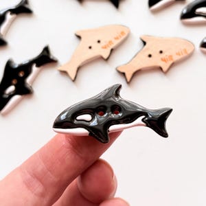 Op de afbeelding: Een verzameling decoratieve knopen in de vorm van orka's. De knopen zijn zwart en wit, met twee gaten om op te naaien. Ook inbegrepen zijn twee lichtbruine knopen in de vorm van een vis met tekst.