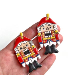 May include: Two decorative nutcracker ornaments. Each ornament is shaped like a soldier with a red hat, red jacket, black boots, and a detailed face. The ornaments are held in a hand against a white background.