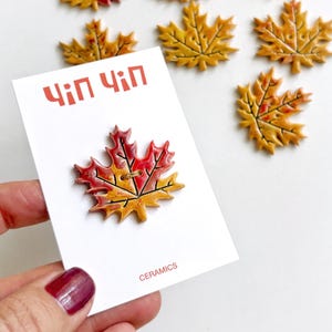 Op de afbeelding: Keramische esdoornbladdecoraties in herfstkleuren rood, geel en bruin. Eén blad is bevestigd aan een witte kaart met rode tekst "Чіп Чіп" en het woord "CERAMICS".