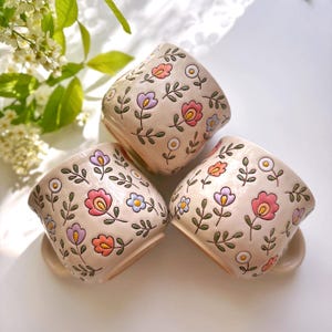 May include: Three beige ceramic mugs with floral designs. Each mug features colorful flowers in pink, purple, and orange, along with green leaves. The mugs are arranged in a close-up shot, showcasing their decorative patterns.