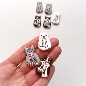 Può includere: Un set di sei bottoni in ceramica a forma di gatto in vari colori, tra cui nero, bianco e grigio. I bottoni presentano un design di gatto dei cartoni animati con occhi neri e un naso rosa.