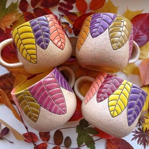 Pode incluir: Quatro canecas de cerâmica com uma base bege salpicada e um padrão de folhas em relevo brilhante em tons de amarelo, roxo, laranja, rosa e verde-oliva. Cada caneca possui uma alça arredondada e um acabamento texturizado e terroso.