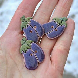May include: Three purple ceramic buttons shaped like aubergines with green leafy tops. The buttons have two holes each.