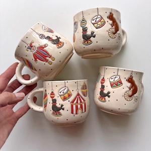 May include: Four ceramic mugs with circus designs. Each mug has a cream base with illustrations of a jester, seals, bears on bicycles, drums, and circus tents. The mugs have a rounded shape with a handle.