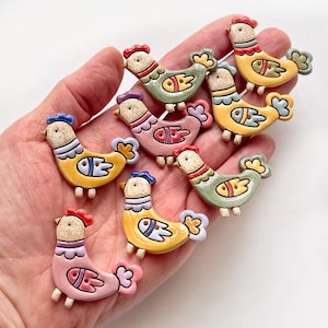 Peut inclure: Un ensemble de sept boutons en céramique en forme de poule dans des tons variés de jaune, rose et vert sauge. Chaque bouton présente une tête beige mouchetée, une crête rouge et une aile stylisée percée de deux trous pour la couture.