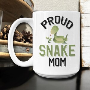 May include: White ceramic coffee mug with a black and green design. The mug features a cartoon snake and the text "Proud Snake Mom".