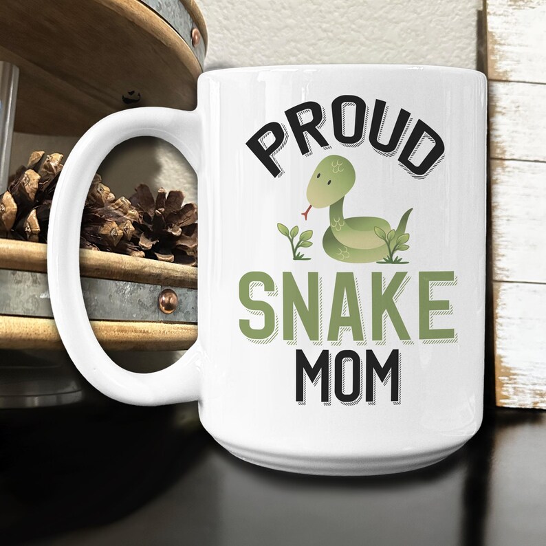 May include: White ceramic coffee mug with a black and green design. The mug features a cartoon snake and the text "Proud Snake Mom".