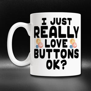May include: White ceramic mug with a black graphic that says "I just really love buttons ok?" with a colorful button design.