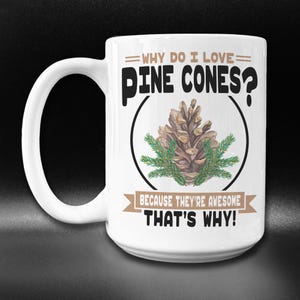 May include: White ceramic coffee mug with a black and brown design. The mug features a pine cone with green needles and the text "WHY DO I LOVE PINE CONES? BECAUSE THEY'RE AWESOME THAT'S WHY!"