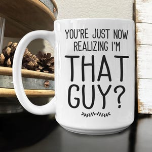 May include: White ceramic coffee mug with black text that reads "You're just now realizing I'm THAT GUY?" with a small leafy design at the bottom.