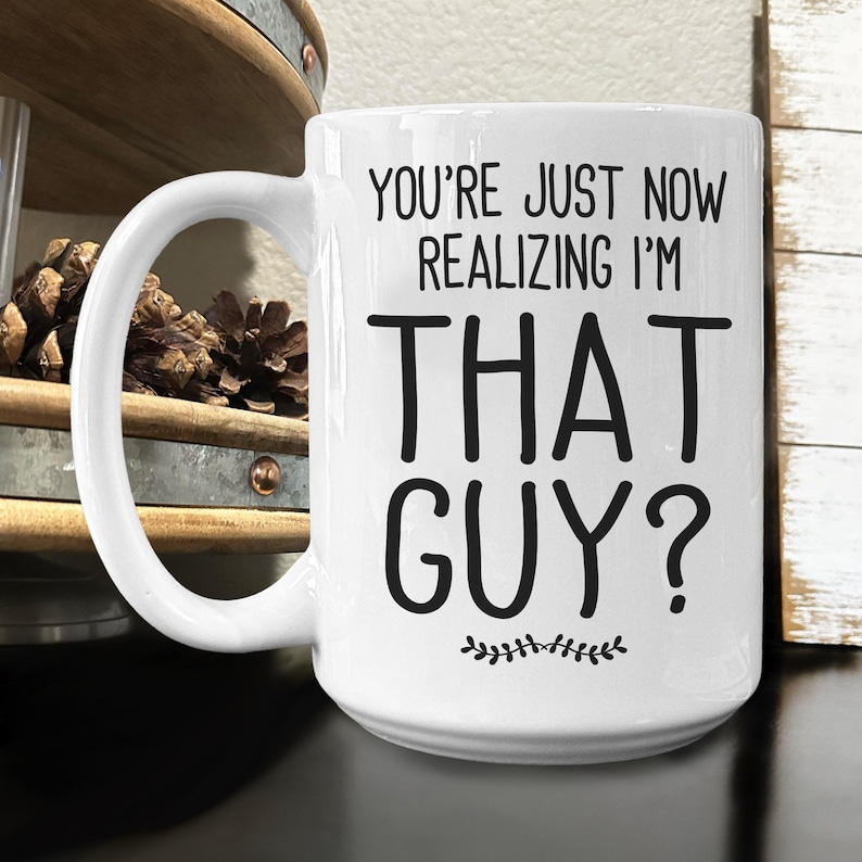 May include: White ceramic coffee mug with black text that reads "You're just now realizing I'm THAT GUY?" with a small leafy design at the bottom.