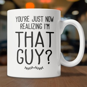 May include: White ceramic coffee mug with black text that reads "You're just now realizing I'm that guy?" with a small decorative leaf design at the bottom.