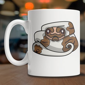 May include: White ceramic coffee mug with a cartoon illustration of a brown and white snake curled up with its head peeking out. The snake has brown eyes and a cute expression.