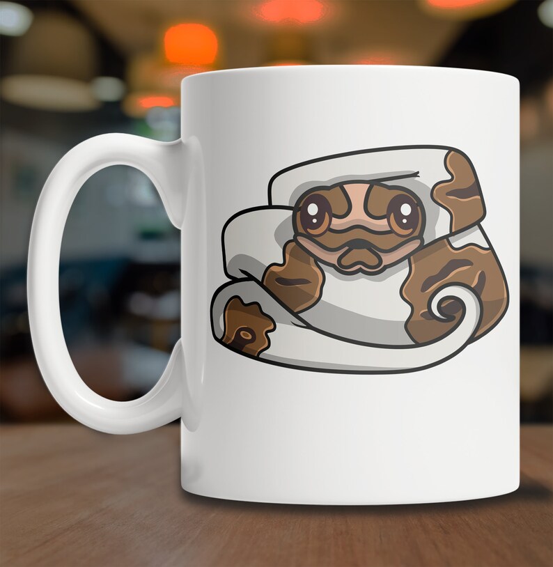 May include: White ceramic coffee mug with a cartoon illustration of a brown and white snake curled up with its head peeking out. The snake has brown eyes and a cute expression.