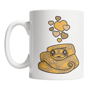 May include: A white ceramic mug with a cartoon illustration of a curled up snake with brown and yellow markings. The snake has large eyes and a small mouth. Above the snake are several small brown and yellow hearts.