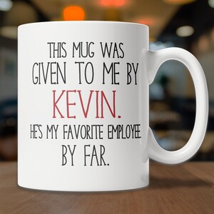 May include: White ceramic coffee mug with black text that reads "This mug was given to me by Kevin. He's my favorite employee. By far."