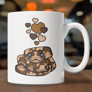 Peut inclure: Une tasse &agrave; caf&eacute; en c&eacute;ramique blanche avec une illustration de serpent brun et beige en dessin anim&eacute; avec des c&oelig;urs flottant au-dessus. Le serpent a de grands yeux et une expression mignonne.