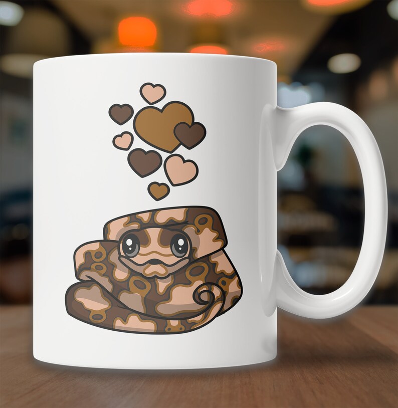 Peut inclure: Une tasse &agrave; caf&eacute; en c&eacute;ramique blanche avec une illustration de serpent brun et beige en dessin anim&eacute; avec des c&oelig;urs flottant au-dessus. Le serpent a de grands yeux et une expression mignonne.