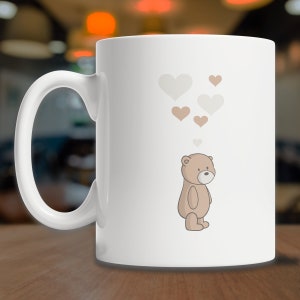 May include: White ceramic mug with a brown teddy bear illustration and a cluster of light brown and white hearts above the bear.
