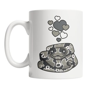 May include: A white ceramic mug with a gray and white cartoon snake with hearts above it. The snake has a cute face and is curled up.