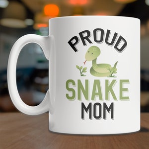 May include: White ceramic coffee mug with a black and green graphic design. The design features a green snake with a red tongue and the text "Proud Snake Mom".