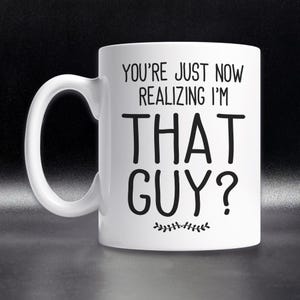 May include: White ceramic coffee mug with black text that reads "You're just now realizing I'm THAT GUY?" with a small decorative leaf design at the bottom.
