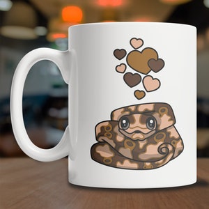 Peut inclure: Une tasse &agrave; caf&eacute; en c&eacute;ramique blanche avec un serpent brun et beige aux gros yeux et un motif de c&oelig;ur au-dessus. Le serpent est enroul&eacute; et a une expression mignonne.