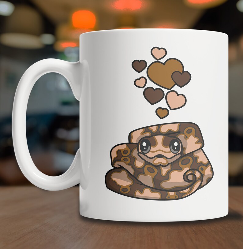 Peut inclure: Une tasse &agrave; caf&eacute; en c&eacute;ramique blanche avec un serpent brun et beige aux gros yeux et un motif de c&oelig;ur au-dessus. Le serpent est enroul&eacute; et a une expression mignonne.