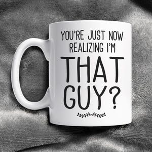 May include: White ceramic coffee mug with black text that reads "You're just now realizing I'm that guy?" with a small decorative line of leaves below the text.