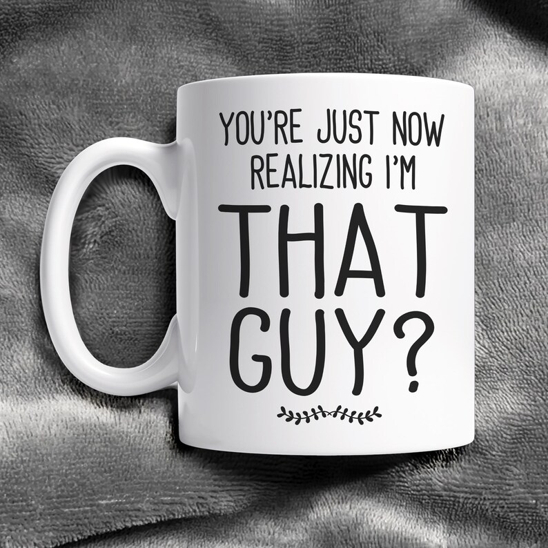 May include: White ceramic coffee mug with black text that reads "You're just now realizing I'm that guy?" with a small decorative line of leaves below the text.