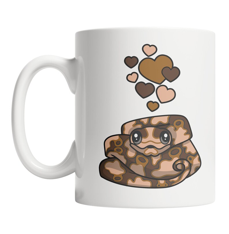Peut inclure: Une tasse en c&eacute;ramique blanche avec un serpent de dessin anim&eacute; brun et beige avec des c&oelig;urs au-dessus. Le serpent a de grands yeux et est enroul&eacute;. Les c&oelig;urs sont bruns et roses.