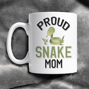 May include: White ceramic coffee mug with a black and green graphic design. The design features a cartoon snake with the text "Proud Snake Mom".