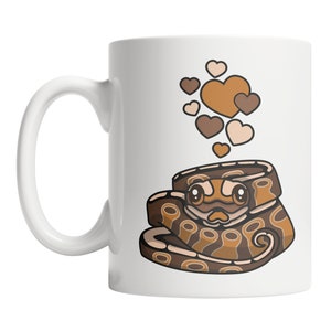 May include: A white ceramic coffee mug with a brown and tan cartoon snake with hearts floating above it. The snake has a cute face with big eyes and a small mouth.