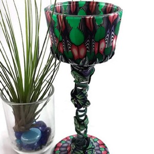 May include: A green and pink patterned glass goblet with a decorative base and stem. The stem is decorated with butterflies and the base is a circular design.