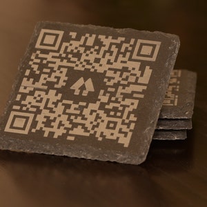 Könnte beinhalten: Ein Satz von vier schwarzen Schieferuntersetzern mit einem weißen QR-Code und einem Logo aus zwei stilisierten Bäumen. Die Untersetzer sind übereinander gestapelt.