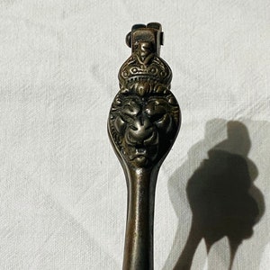 Op de afbeelding: Antieke notenkraker in bronskleur met een leeuwenkopontwerp. De notenkraker heeft een gedetailleerd leeuwen gezicht met een kroon erboven. De handgreep is lang en slank, en het algehele ontwerp is sierlijk en decoratief.