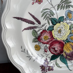 Könnte beinhalten: Weißer Keramikteller mit gewelltem Rand, verziert mit einem Blumendesign in Rot-, Rosa-, Gelb- und Lilatönen. Die Blumen werden von grünen Blättern und zarten Stielen begleitet. Der Teller hat eine glänzende Oberfläche.