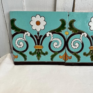 May include: Two decorative ceramic tiles with a turquoise background. Each tile features a white daisy, brown and white scrollwork, and green and orange accents. The tiles have a dark brown border.