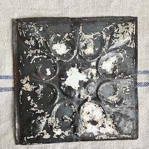 May include: A vintage tin ceiling tile with a decorative, embossed pattern of swirling circles and leaves. The tile is painted in a distressed white and grey finish.