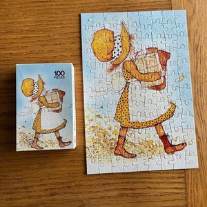 Puede incluir: Un rompecabezas de 100 piezas con una niña con un vestido y un sombrero amarillos que lleva un paquete grande. El rompecabezas está sobre un fondo azul con un borde blanco.