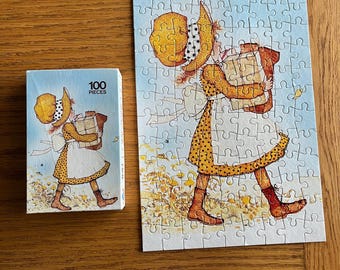 May include: A 100 piece jigsaw puzzle featuring a girl in a yellow dress and hat carrying a large package. The puzzle is on a blue background with a white border.