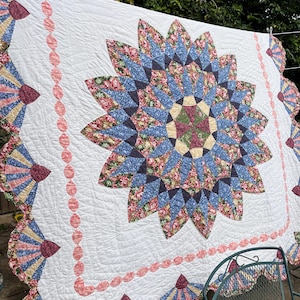 May include: A white quilt featuring a large central star pattern composed of blue floral and solid dark fabric segments. The quilt is bordered by a pink circular chain motif and scalloped fan-shaped edges with floral and striped fabric patterns.