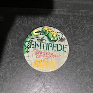 May include: A round Centipede Atari button featuring a green and yellow centipede illustration. The button has the text "CENTIPEDE" in green, "New and only from" in red, and "ATARI" in orange.