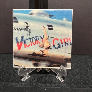 May include: A ceramic tile with a vintage pin-up style image of a woman in a swimsuit standing on a plane with the words "Victory Girl" in red and blue letters.