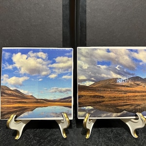 May include: Two square ceramic coasters with a landscape image of a lake and mountains reflected in the water. The sky is blue with white clouds. The coasters are on white stands with gold accents.