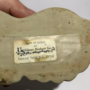 May include: A white ceramic figurine with a label that reads "Made in Taiwan for U.S. Leisure Products Inc. Central Islip, N.Y. 11722"